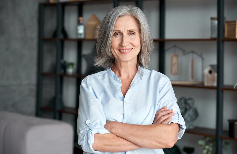 A woman with gray hair and a blue shirt stands smiling with arms crossed in front of a bookshelf.