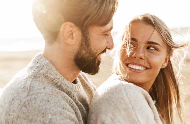 A couple wearing sweaters smiles at each other while sitting outdoors on a sunny day.